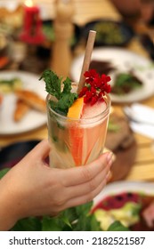 Glass Of Refreshing Lemonade With Grapefruit Slices, Decorated With Leaves And Flowers, With Eco Paper Straw. Summer Drink, Cocktail, In A Tall Glass In A Hand, With Blurred Background.