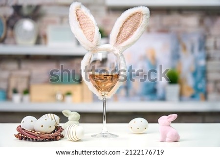 Glass of rose or red wine with bunny ears and Easter decorations, colorful eggs on white table, on bright background at home. Copy space for text.