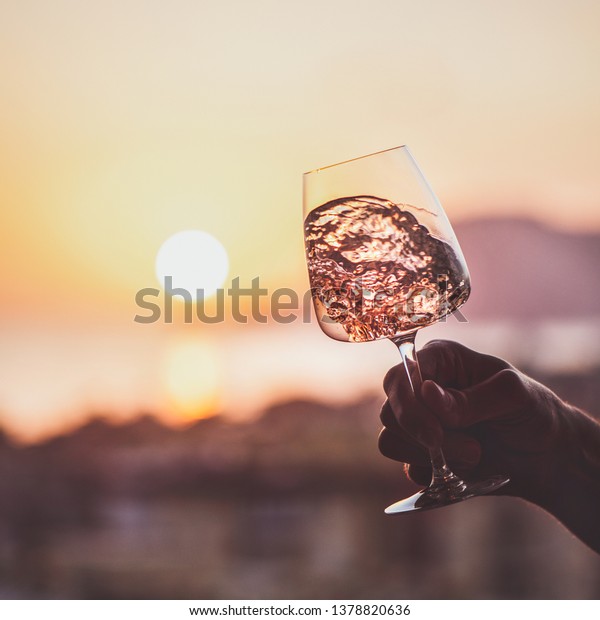 Glass of rose wine in mans hand with sea and\
sunset at background, close-up, square crop. Summer evening relaxed\
mood concept