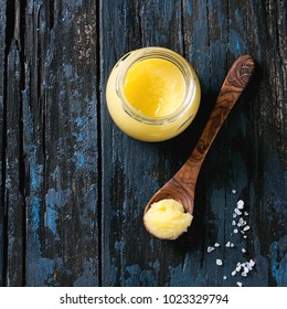 Glass of salted drawn ghee butter standing with wood spoon and sea salt over old dark blue wooden background. Healthy eating. Top view, copy space. Square image