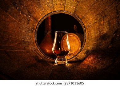 A glass of scotch whiskey in old oak barrel. Copper alambic and small barrel on background. Traditional alcohol distillery concept - Shutterstock ID 2371011605