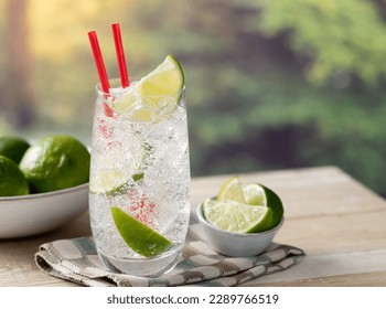 Glass of sparkling cold water with lime wedges and ice outdoors on a wooden table with summer background