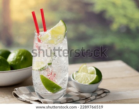 Glass of sparkling cold water with lime wedges and ice outdoors on a wooden table with summer background