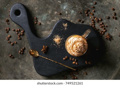 Glass of spicy pumpkin latte with whipped cream and cinnamon standing on black serving board. Coffee beans and spices above. Dark background. Top view, space.