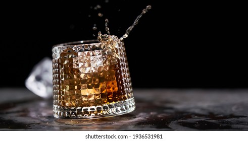 Glass of splashing whiskey with icecube on stone table, on black background. Whiskey splash concept, copy space for text.