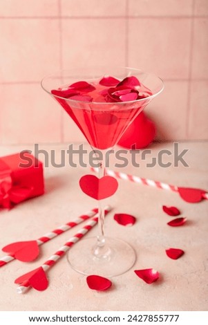 Glass of tasty cocktail with rose petals and gift on grunge table against beige tile background. Valentine's Day celebration