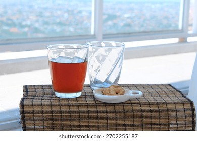 A glass of tea with two empty glasses and a small plate of snacks on a bright windowsill, creating a simple and relaxed refreshment scene.