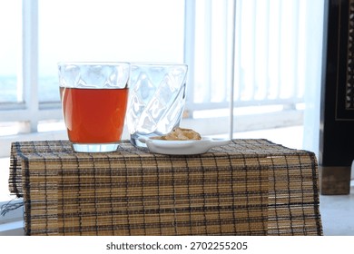 A glass of tea with two empty glasses and a small plate of snacks on a bright windowsill, creating a simple and relaxed refreshment scene.