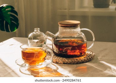 Glass teapot with brewed tea, cup of tea and lime under warm sunlight on the table with green leaf in the background - Powered by Shutterstock