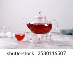 Glass teapot with deep red herbal brew on a clear stand, accompanied by a matching glass teacup, set against a soft, muted background.