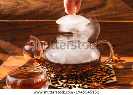 the glass teapot in which the tea is poured stands on a wooden background. a woman's hand removes the lid from the kettle
