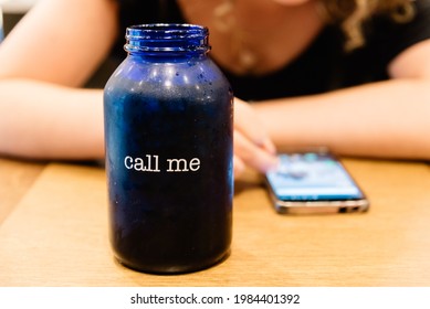 Glass With The Text Call Me On Background Of Woman Texting On Phone Waiting For Answer Or Phone Call. Focus On Glass. Frustrated Social Outcast Or Loner Sitting Alone At Coffeeshop Table