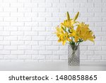 Glass vase with yellow lily flowers on marble table near white brick wall, space for text