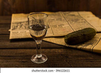Glass of vodka with newspaper and pickled cucumber on an old wooden table. Angle view, shallow depth of field, focus on the glass of vodka