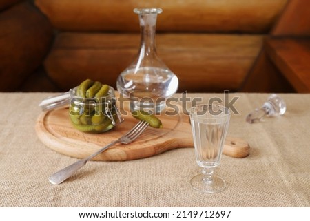 A glass of vodka is on the table next to pickles and a decanter on the background of a log wall. 