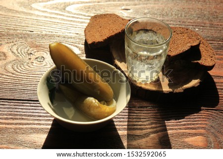 A glass of vodka and pickled cucumber with brown bread on the table. Shadow of food on a wooden table.