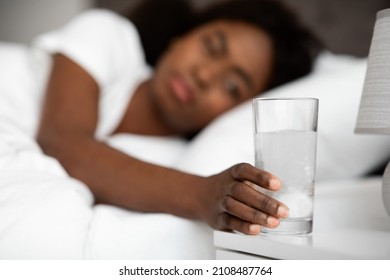 Glass Of Water With Effervescent Tablet On Bedside, Sick African American Young Woman Laying In Bed And Reaching Medicine, Feeling Bad, Experiencing Hangover In The Morning, Blurred Background