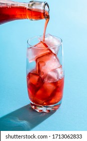 Glass Of Water With Ice Cubes And Strawberry Syrup. Pouring Syrup From A Glass Bottle To A Glass Full Of Ice Cubes. Drink Preparation In Bright Light On A Blue Colored Background