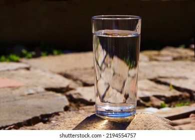 A Glass Of Water Stands On A Gray Broken Paving Slab.