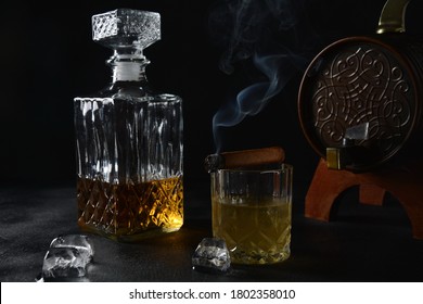 Glass Of The Whiskey With Ice Cubes And A Square Decanter, And Cigar On A Black  Background.