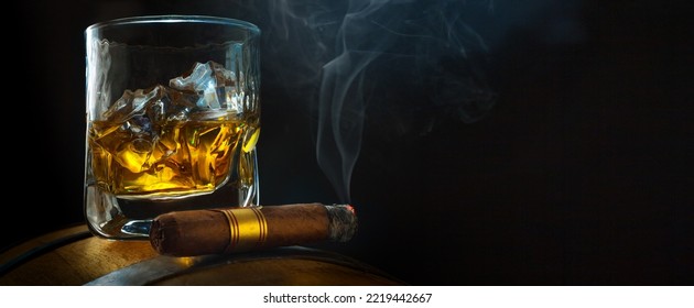 A Glass Of Whiskey With Ice On A Oak Barrel, A Steaming Cuban Cigar  In A Smoky Atmosphere Of A Night Bar. Men's Club Banner Idea. Copy Space For Text