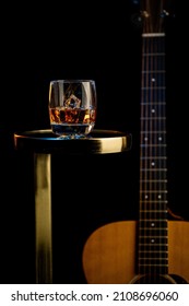 A Glass Of Whiskey With Ice Sits On A Nightstand Next To An Acoustic Guitar