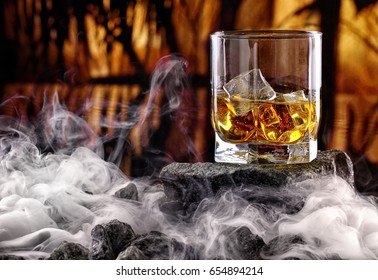 Glass of whiskey and ice.Creative photo glass of whiskey on stone with fog and sunset background.Copy space.Advertising shot