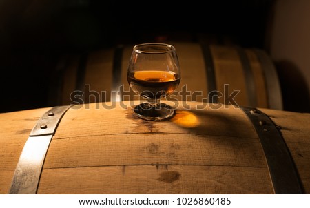 A glass of whiskey sits atop of a whiskey barrel that is aging product. The look of the photo has a vintage feel with a modern spirit.