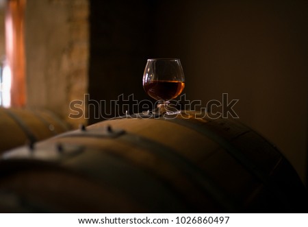 A glass of whiskey sits atop of a whiskey barrel that is aging product. The look of the photo has a vintage feel with a modern spirit.