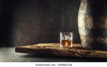Vidrio de whisky de coñac o borbón en vidrio ornamental junto a un barril de madera de vinatge sobre una madera rústica y fondo oscuro.