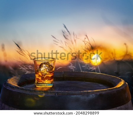 Glass of whisky with ice cubes on old wooden cask. Beautiful sunset at the background.