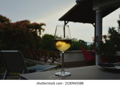 Glass Of White Wine, Misted From A Golden Drink, Stands On A Table In The Garden Against The Backdrop Of A Free Space Sunset. Glass Isolated, Marco Photo, Garden Full Of Flowers, Hungary, Lake Balaton