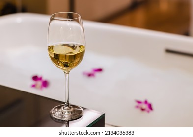 A Glass Of White Wine Puts On Table Beside Bathtub That Have White Bubble And Orchid Flowers. Relaxing At Home Concept.