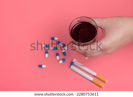 A glass of wine held in the hand on a pink background next to cigarettes and pills