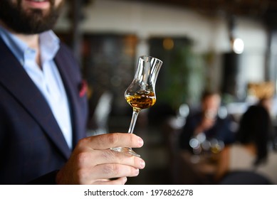 A Glass With Wiskey In Hand Of Man With Blurred Background. Concept Of Whiskey Tasting.