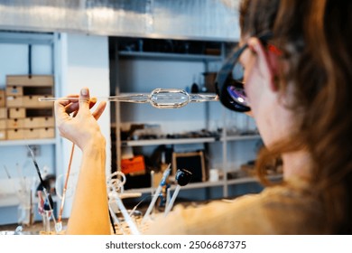 A glassblower meticulously inspects a delicate piece of glasswork, holding it up to the light in a well-lit workshop. - Powered by Shutterstock