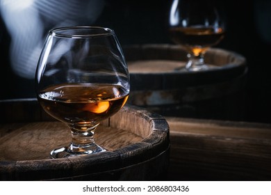 Glasses of cognac of amber color stand on barrels of cognac in wine cellar illuminated by soft light. Brandy in glasses on wooden barrel of amber-colored alcohol.
