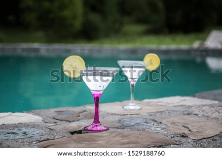 Glasses of martini cocktail at swimming pool with forest trees on background. Selective focus. Season and holidays concept.