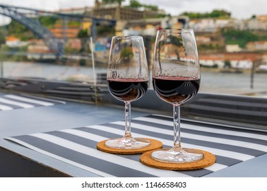 Glasses Of Port Wine At An Outdoor Restaurant With The Douro River And Dom Luis I Bridge Blurred In The Background In Porto, Portugal