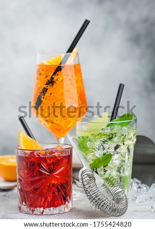 Glasses of spritz,mojito and negroni cocktails with ice cubes and lime and orange slices with mint leaf and black straw on light table  background with strainer. 