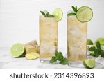 Glasses of tasty ginger ale with ice cubes and ingredients on white marble table