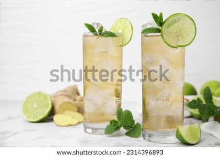 Glasses of tasty ginger ale with ice cubes and ingredients on white marble table