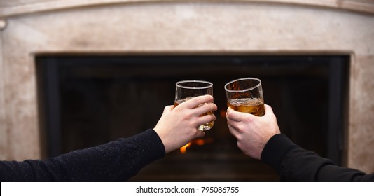 Glasses With Whisky In Male Hands. Hands Clinking Whisky Glasses On Fireplace Background. Male Hands With Drinks In Glasses At Home By Fireplace. Men Cheers With Whisky In Glasses Near Fireplace