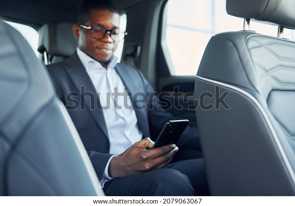 In glasses. Young african american\
businessman in black suit is in the\
automobile.