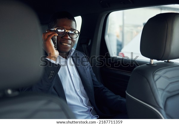In glasses. Young african american\
businessman in black suit is in the\
automobile.