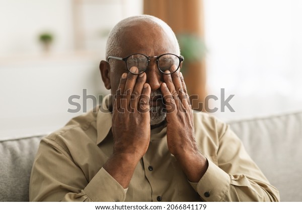 Glaucoma.\
Senior African American Man Rubbing Tired Eyes Wearing Eyeglasses\
Having Poor Eyesight Sitting On Couch At Home. Ophtalmology\
Diseases, Health Problems In Older Age\
Concept