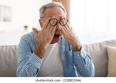 Glaucoma. Senior Man Rubbing Tired Eyes Wearing Eyeglasses Having Poor Eyesight Feeling Suffering From Eyestrain Sitting On Couch At Home. Ophtalmology Diseases, Health Problems In Older Age Concept