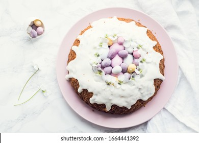 Glazed easter cake decorated with flowers and mini chocolate eggs candy on white marble background. Happy Easter Holidays. Top view. Copy space.