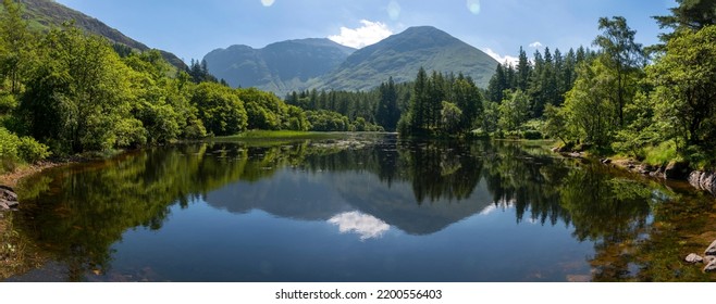 Glen Coe In The Lochaber Area Of The Scottish Highlands.Distant Mountains Reflected In The Still Waters Of A Calm,within A Forest Lake On A Hot Summer Afternoon.