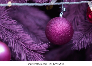 Glittering purple bauble hanging on a vibrant purple Christmas tree branch, adorned with soft warm fairy lights - Powered by Shutterstock